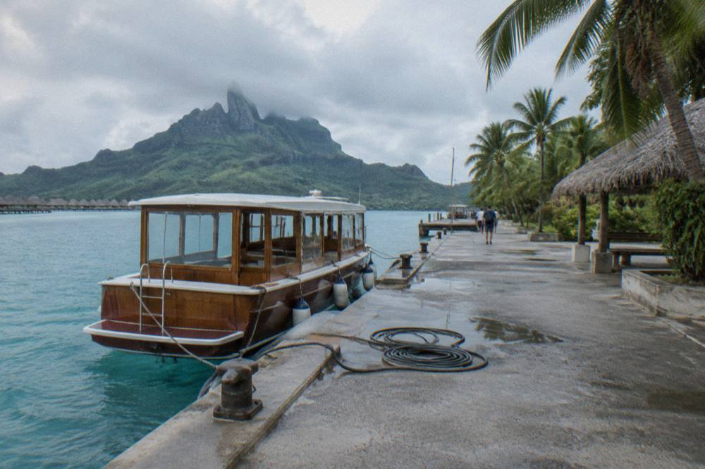 Wooden tender boat tied at a small concrete pier beside palm trees and calm blue water