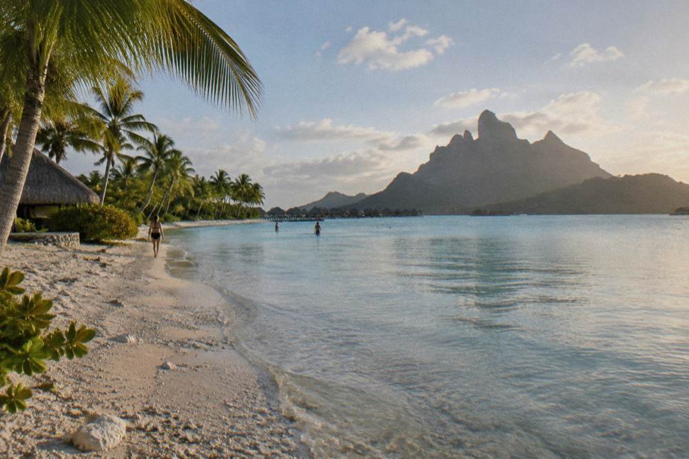 A palm-fringed motu beach with calm shallow water and a wooded island peak beyond