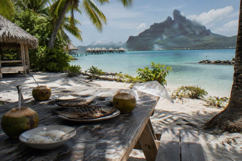 Wooden picnic table set with lunch on a sandy motu beside calm lagoon water