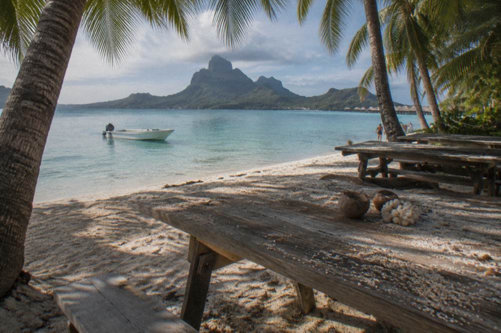 A small boat moored beside a palm-fringed motu with lunch tables set under shade