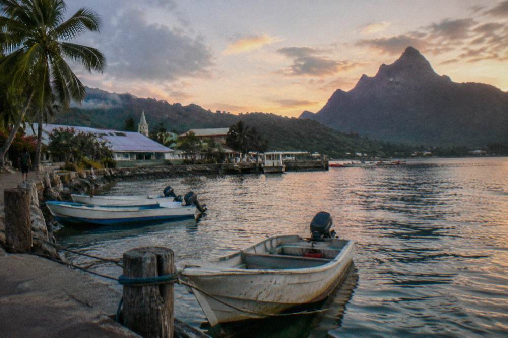 Small waterfront town along a quay with boats moored in calm water and green hills behind