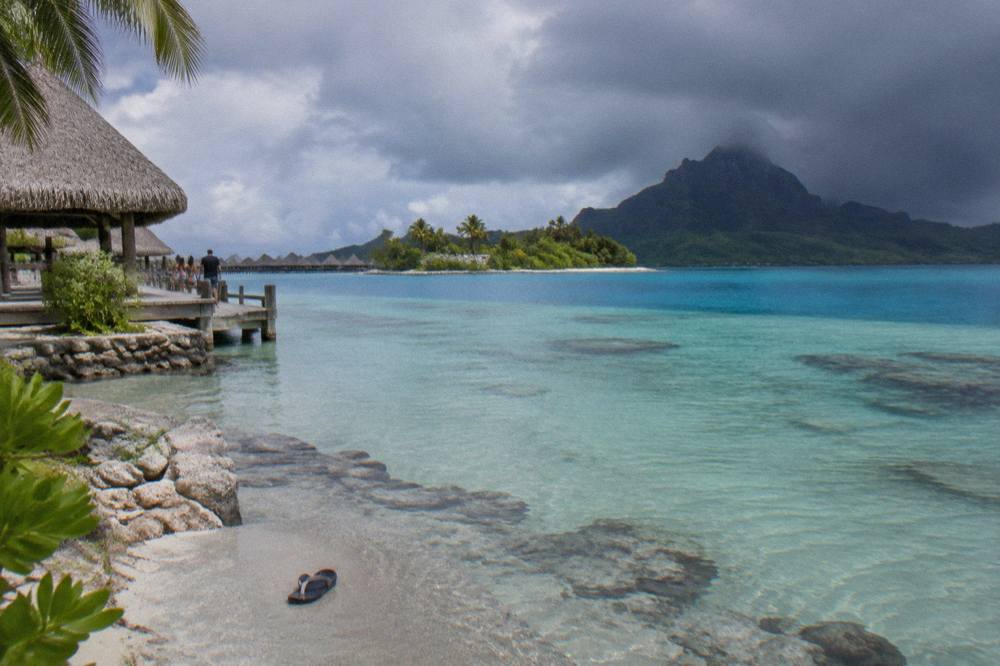 Calm shallow lagoon water in shades of blue beside a green motu shoreline