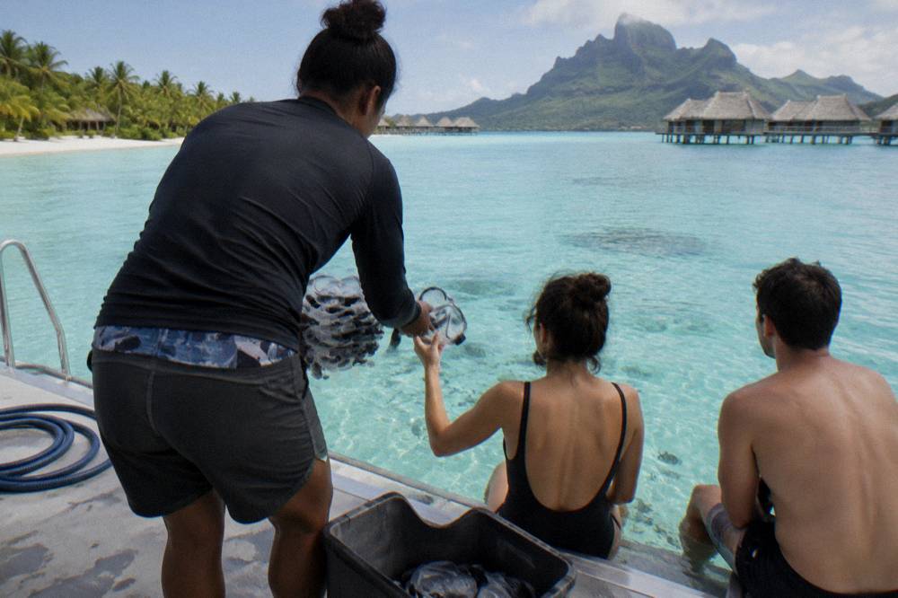 A guide handing snorkel masks to guests on a boat deck over clear water