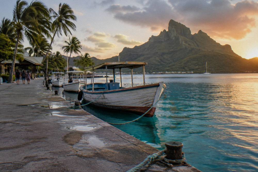 Wooden boats moored beside a quiet quay with palm trees and green cliffs behind