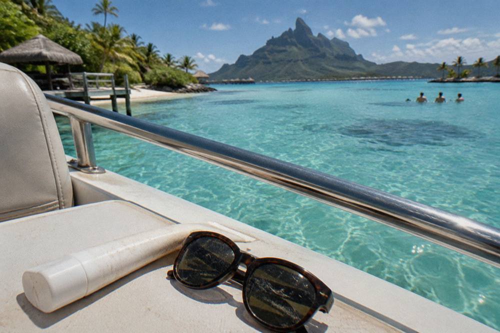 A tube of mineral sunscreen and sunglasses on a boat deck with lagoon water beyond